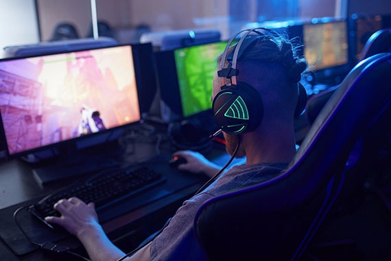Rear view of young gamer wearing gaming headphones with backlight and playing in computer video game on computer in dark computer class