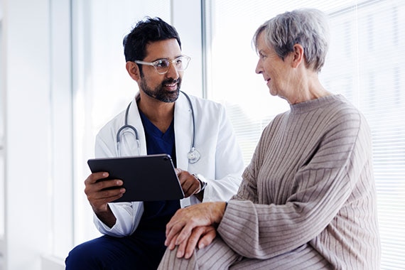 Healthcare consultation in a clinical setting with a healthcare professional using a tablet while meeting with an older patient.