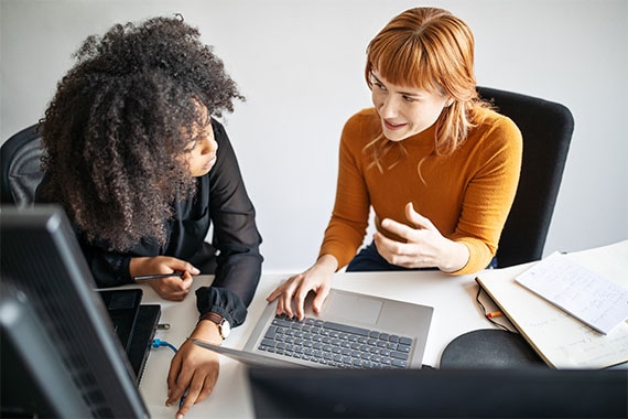 Two people review information on a laptop at a desk, representing device selection, product comparison, and online purchasing workflows.