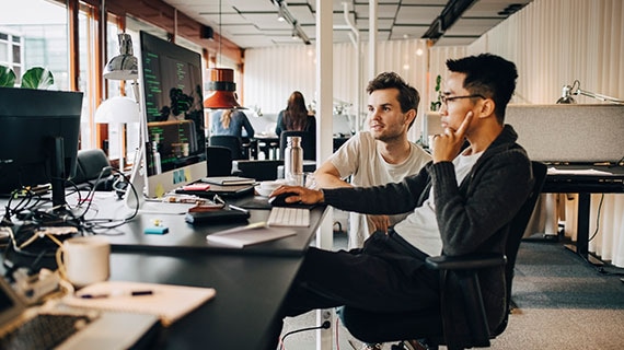 Two software developers reviewing code and system data on a workstation in a modern office environment, representing the development of a compliance-focused audit log endpoint with traceable activity tracking and secure backend services.