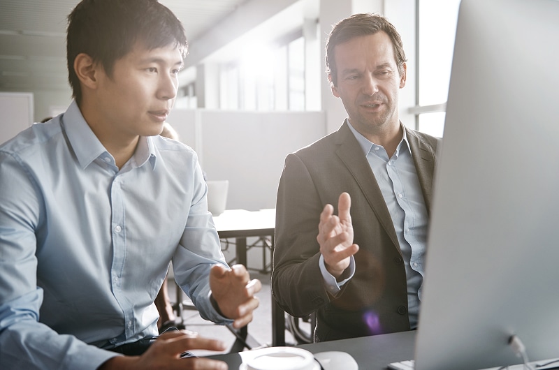Two businessmen talking while looking at a desktop monitor in a bright office setting.