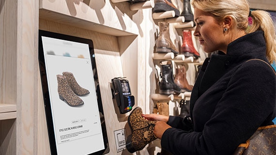 Shopper using an interactive Elo touchscreen kiosk to browse and purchase shoes in a retail store, showcasing versatile self-service and endless aisle solutions.