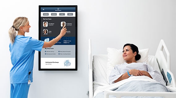 Nurse using an interactive Elo touchscreen display in a hospital room to enhance patient care, with a patient resting in bed.