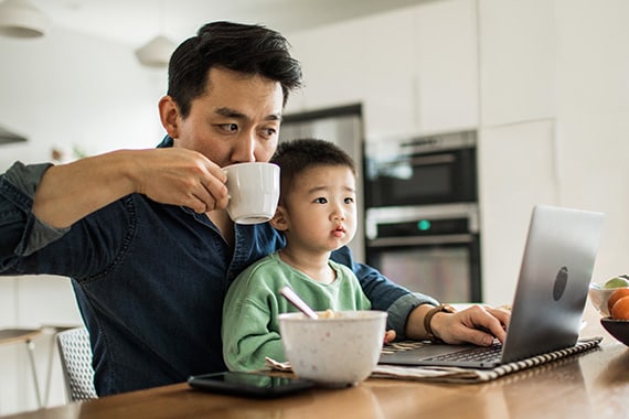 man remotely working at home on laptop in kitchen with child in lap