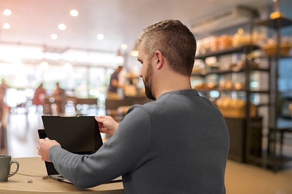 Man using a laptop with a privacy screen filter in a modern café, demonstrating Kensington's Eyesafe Blue Light Technology for eye health and screen privacy.
