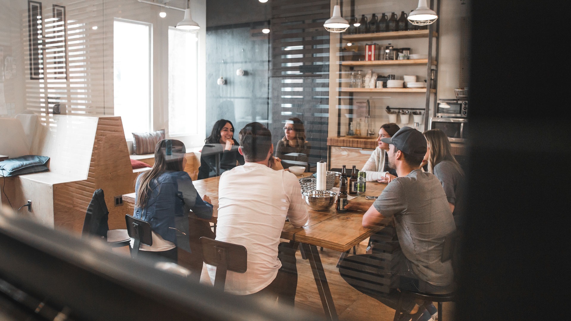 Overlooking from behind a glass wall into a kitchen area where people are sitting and having a discussion.
