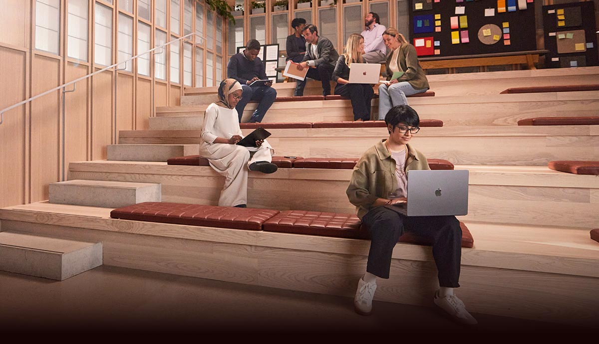 Woman working on a MacBook in a communal space with other other employees.