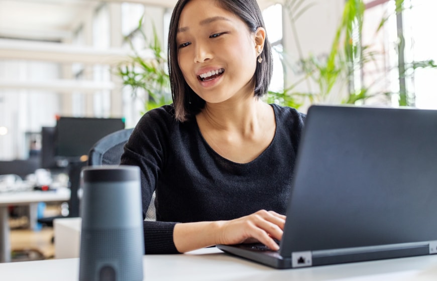 Close up image of a woman at her desk speaking to a smart speaker.