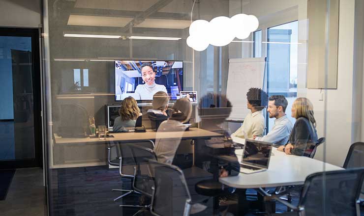 Coworkers gather at a boardroom table as they watch another coworker present on a projected screen in front of them