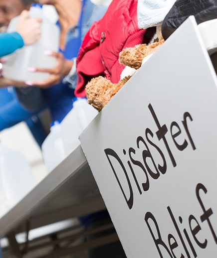 A white sign reads “Disaster Relief” in black letters as relief items are exchanged in a blurred background