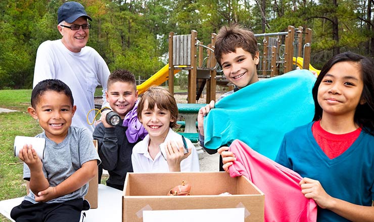 Group of children hold objects from a box in front of them as an adult stands behind them smiling