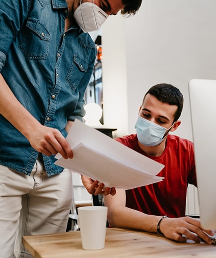  masked coworker shows another masked coworker, seated at a computer, a series of papers