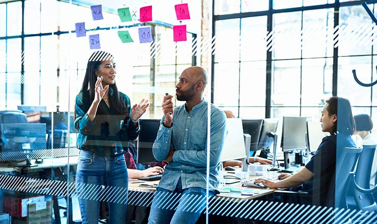Two coworkers with markers collaborate on a clear writing board in an office setting