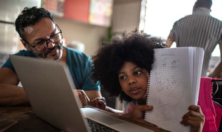 Adult looks on as a child shows the work she has done on her notebook to the laptop’s camera