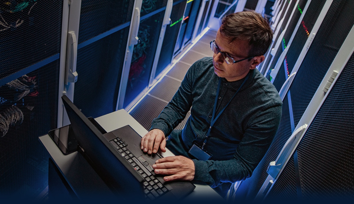IT specialist working on a laptop in a server room surrounded by network racks.