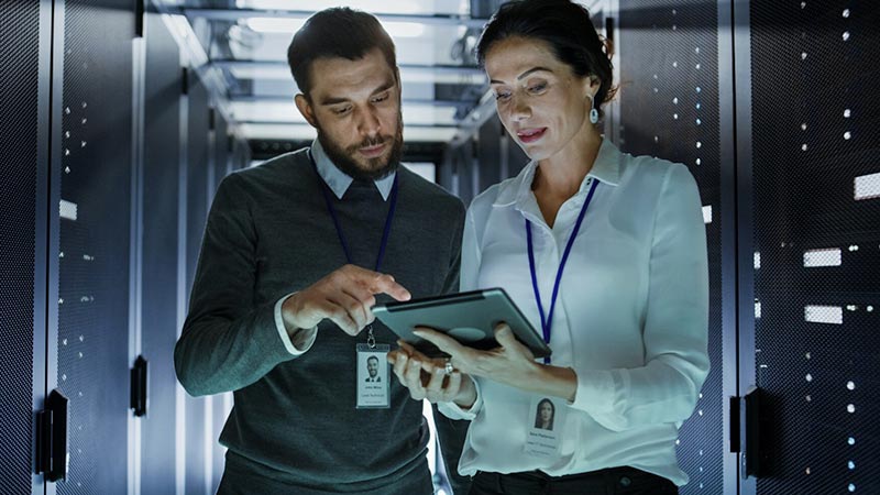 Two IT Engineers Walking in Data Center / Server Room, Talking, Running Diagnostics with Tablet Computer.