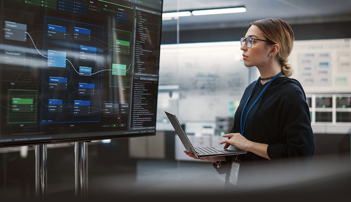 Woman analyzing data flow diagrams on a large monitor while working on a laptop.
