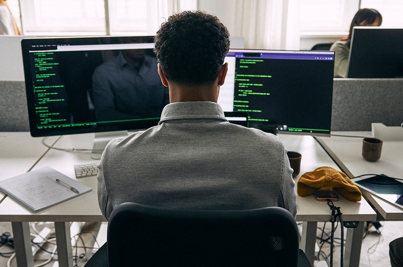Back view of a programmer coding on dual monitors in a modern office workspace.