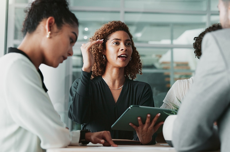 Woman holding a tablet and speaking during a team discussion in a glass-walled meeting room.