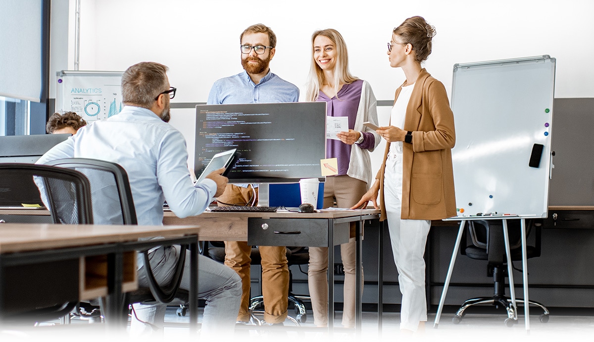 Team discussing project around a desk in a modern office with monitors and notes.