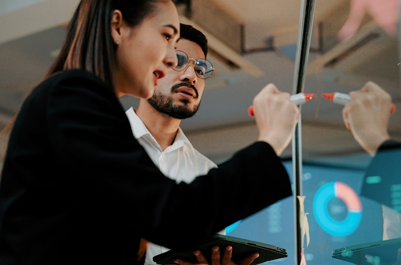 Two colleagues brainstorming and drawing on a transparent board in front of data screens.