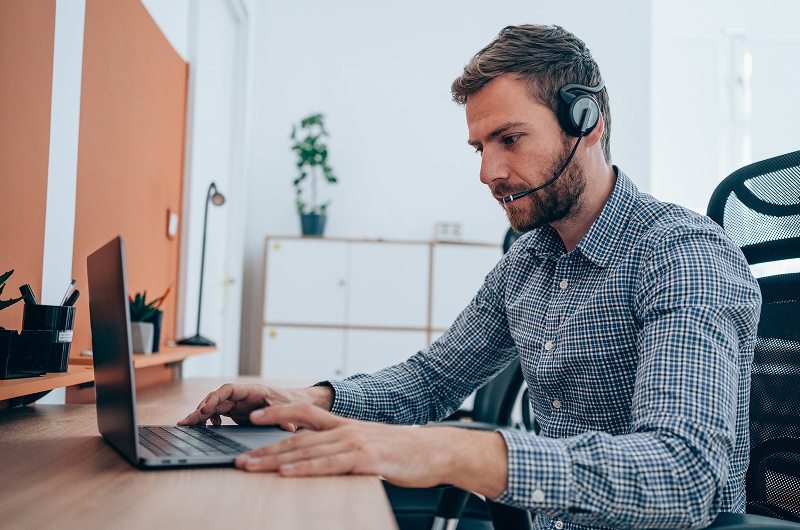 Customer support agent with a headset typing on a laptop in a bright office workspace.