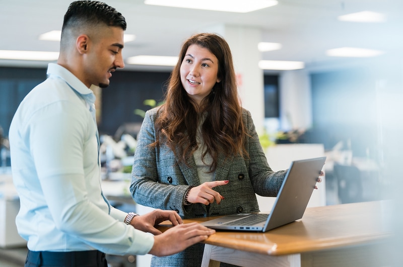 Two colleagues standing at a desk with a laptop, discussing work in a modern office setting.