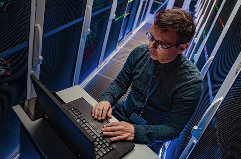 IT specialist working on a laptop in a server room surrounded by network racks.