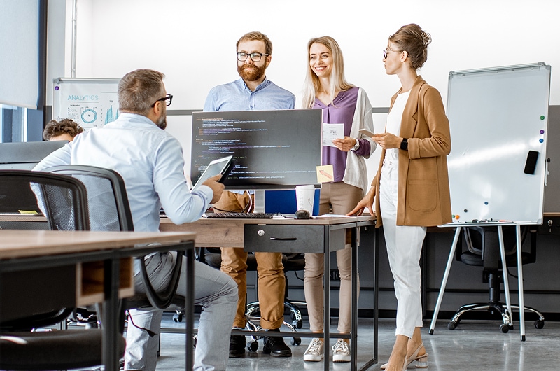 Team of developers discussing code around a desk in a modern office with monitors and notes.