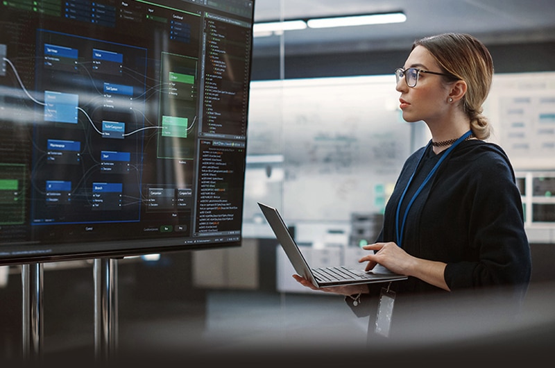 Woman analyzing data flow diagrams on a large monitor while working on a laptop.