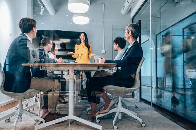 Business team in a modern glass-walled conference room listening to a presenter, symbolizing AI-driven collaboration and strategic decision-making to accelerate business outcomes.