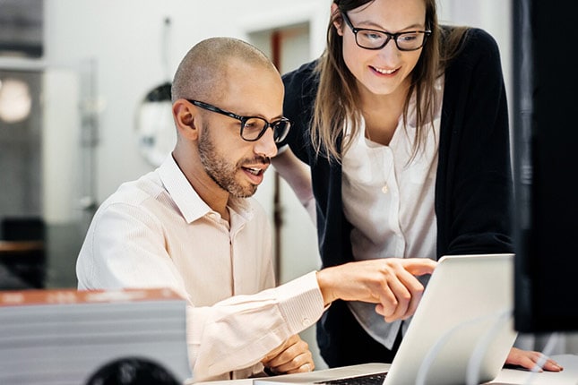 Businessman is sitting in front of a notebook in a modern office room, explaining something to a female colleague standing next to him. He's pointing at the screen while the woman is happily listening to him.