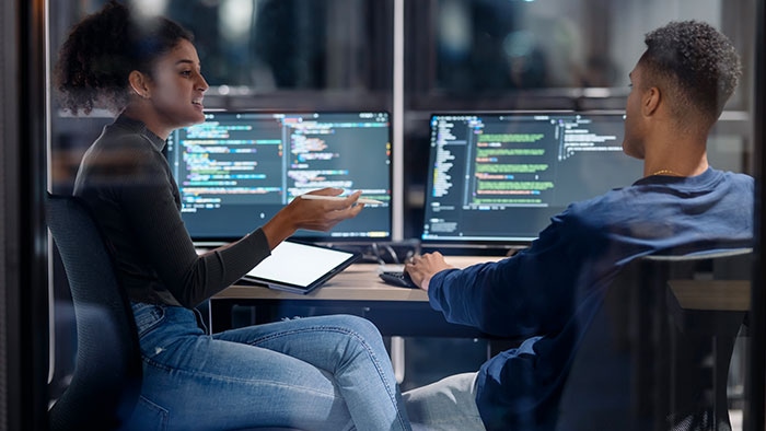 Two professionals collaborate at a workstation with code on multiple monitors, illustrating data governance framework implementation and data quality management.