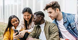 A group of four young people sit on a curb as they gather around a young woman’s phone screen