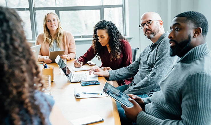 Five coworkers seated at a table in an office are engaged in a meeting, writing down notes and referencing their respective laptops