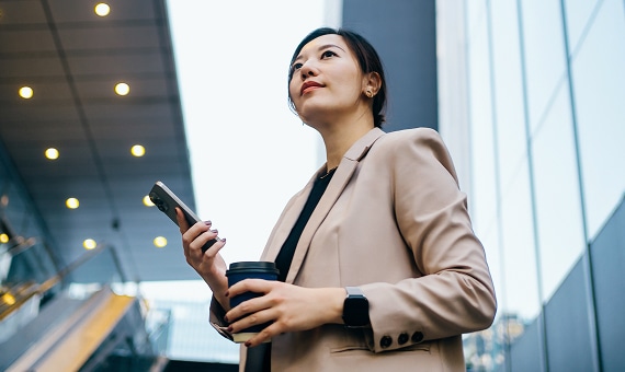 Professional holding a smartphone and coffee while standing outside a modern office building.
