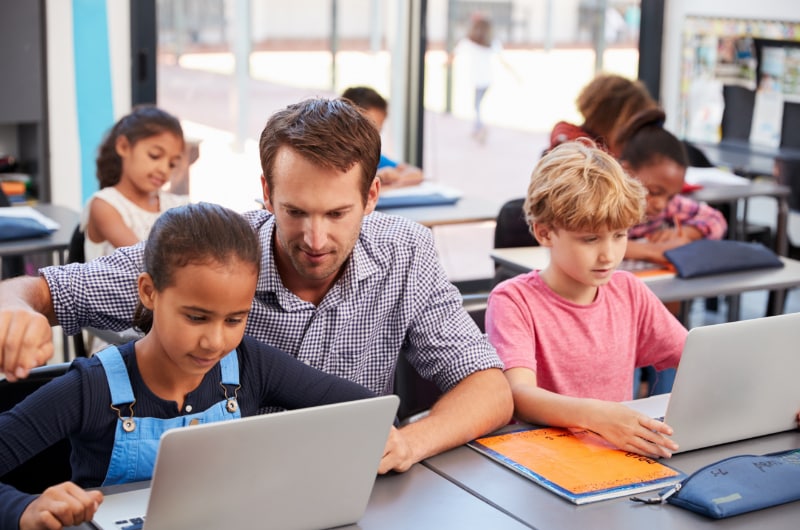 A male teacher helps a young student with her computer.