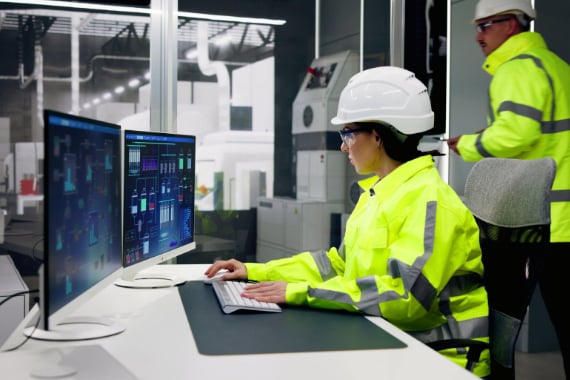 A woman in safety gear works on a computer at a desk in a manufacturing plant office.