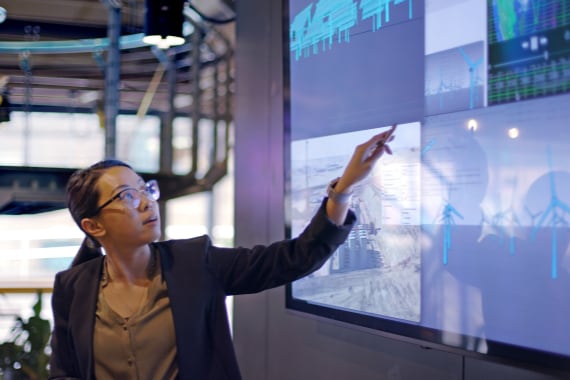 A female professional stands before a large monitor on the wall, pointing to data about wind power operations.