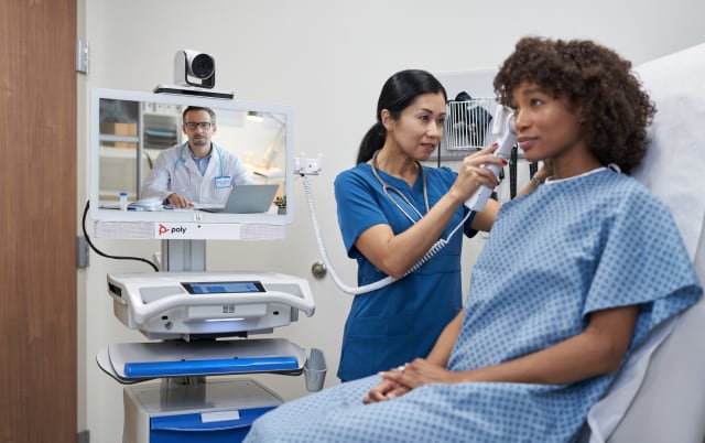 Image of a nurse checking the temperature of a patient with the doctor video call behind through a Poly device 