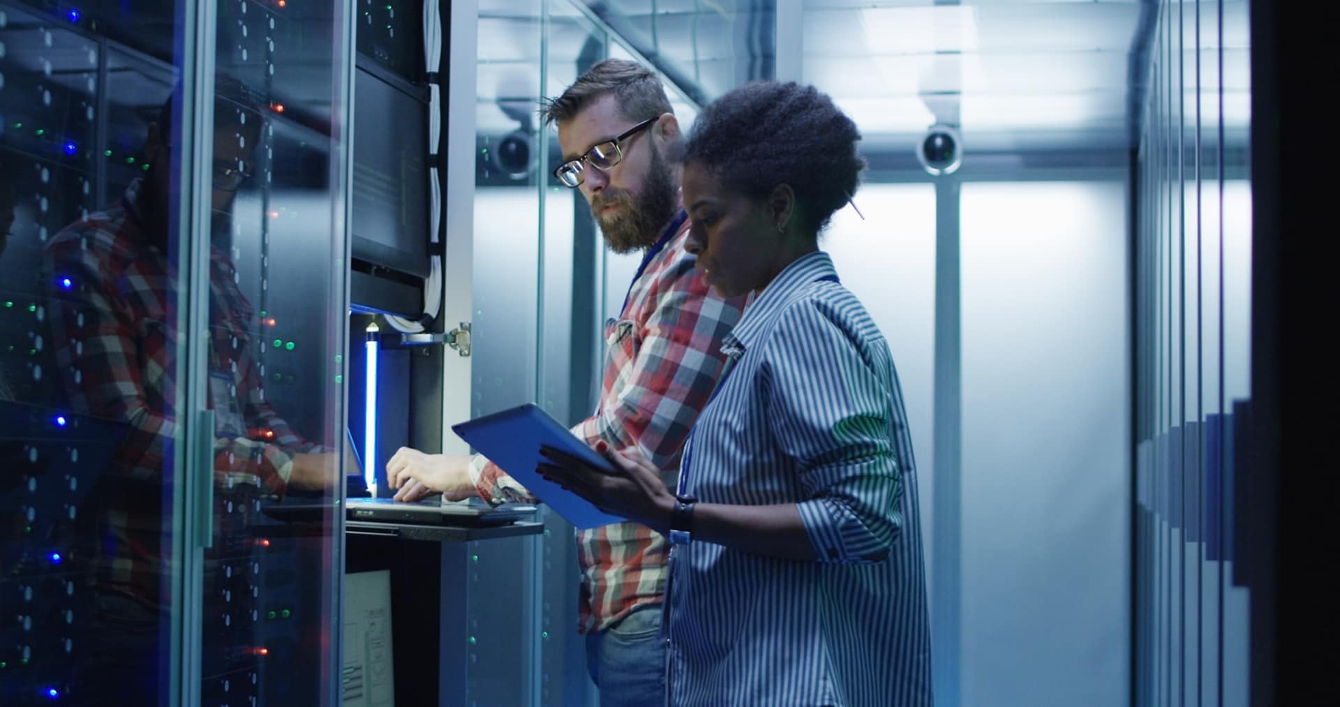 Modern multiethnic man and woman with tablet using laptop in server room while checking servers