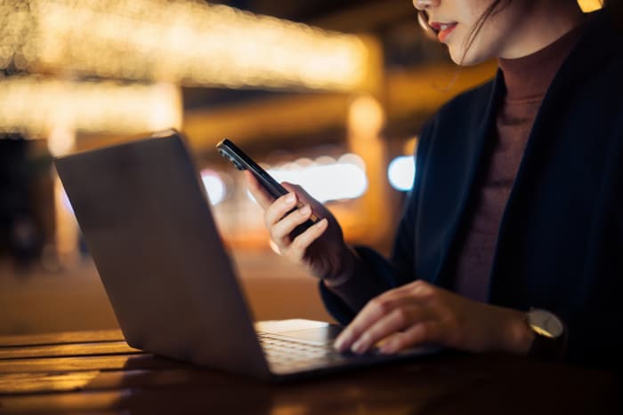 Close up shot of professional young Asian businesswoman using smartphone while working on laptop, sitting outdoors in a side walk cafe at night. Work remotely. Self-employment and freelancer working in the city, Business on the go