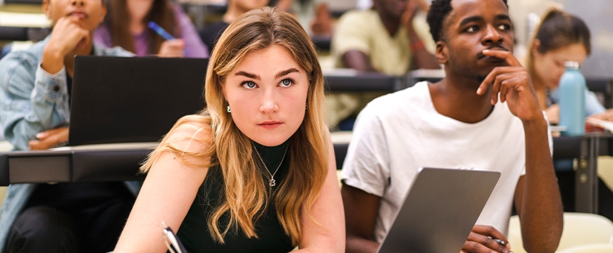 Young female student writing in book while sitting by male friend at university classroom