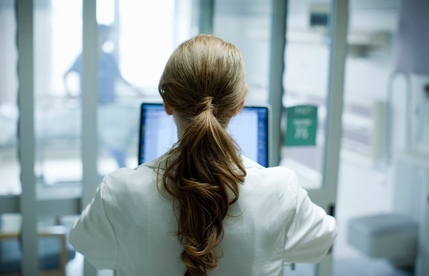 Woman doctor with long red hair using laptop in hospital, photographed from behind