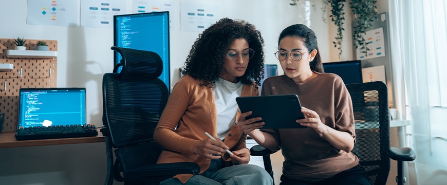 Women in tech collaborate on UI review with tablet and coding monitors in background