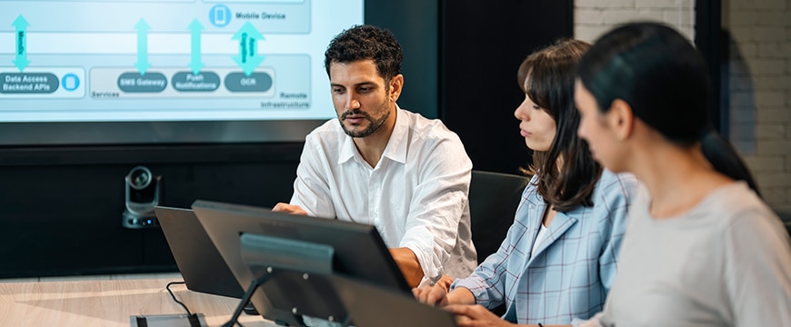 Three colleagues sit at a conference table working on laptops, with a large screen in the background displaying a technical diagram featuring data access, SMS gateway, and mobile services components.