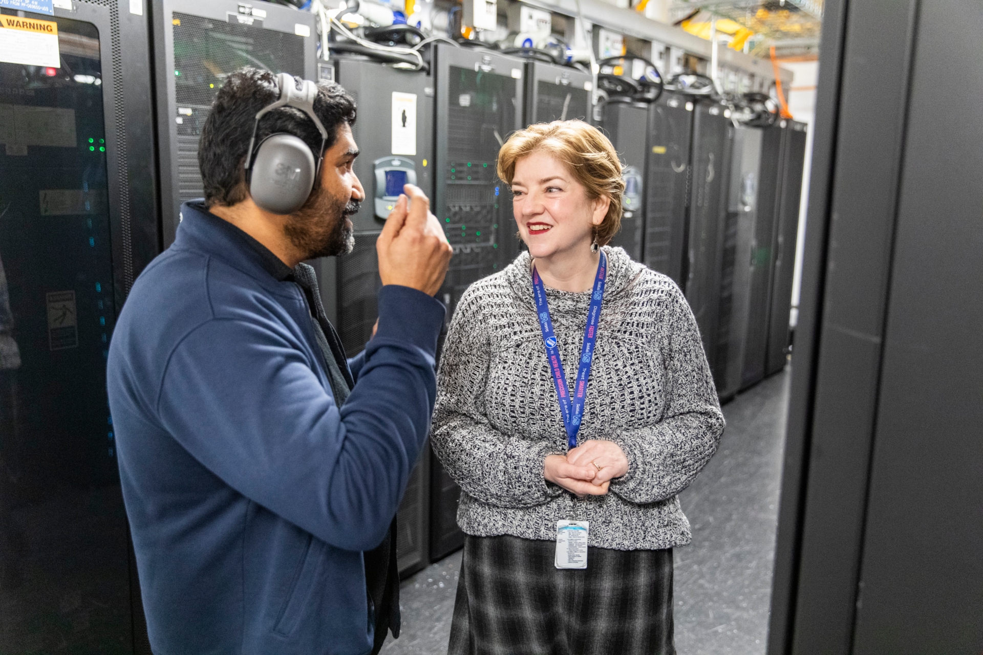 Two IT professionals stand in a data center corridor, talking beside tall server racks. The man on the left wears hearing protection headphones and holds a small device while speaking. The woman on the right, wearing a patterned sweater and plaid skirt, listens and smiles, with an ID badge and lanyard visible. The environment is filled with computer cabinets, cables, and indicator lights, suggesting a technical workplace setting.