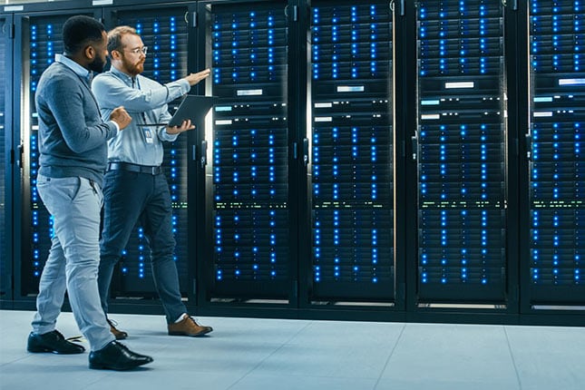 IT Technician with a Laptop Computer and Black Male Engineer Colleague are Talking in Data Center while Walking Next to Server Racks. Running Diagnostics or Doing Maintenance Work.