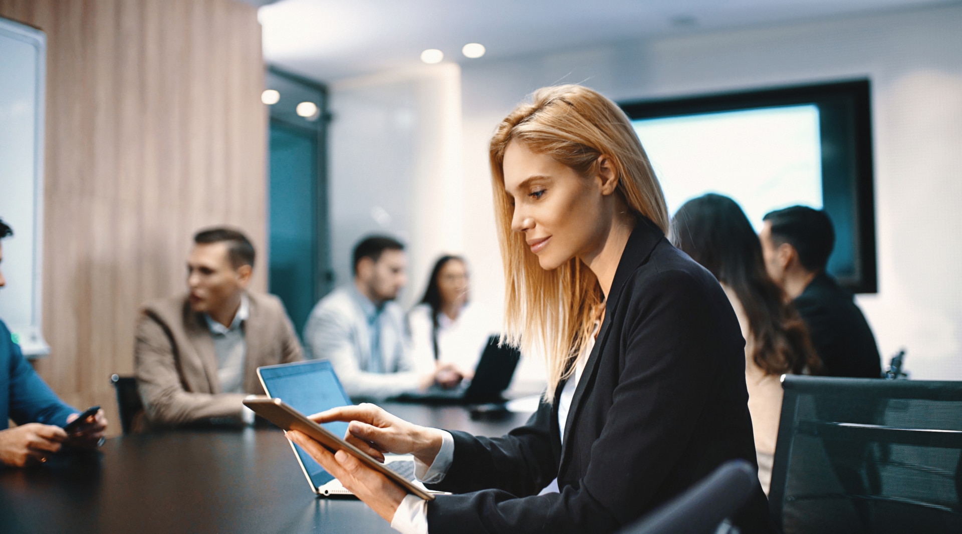 Closeup of a board room meeting at a business company, usual scene at any modern company. There are four men and three women, one of the women is sitting in foreground sideways to the camera and using a digital tablet.