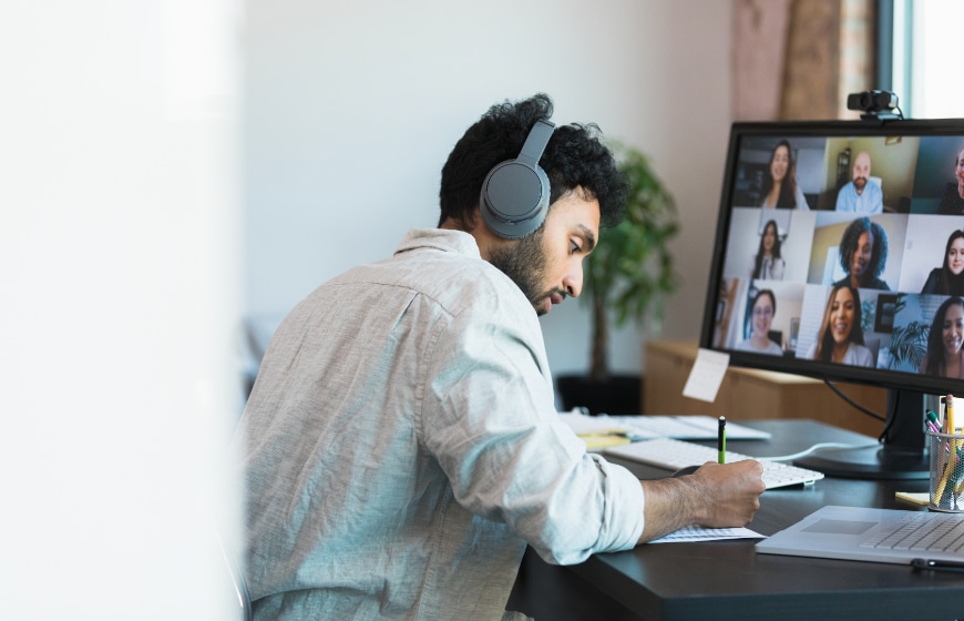 Image of a man with sitting at desk with computer screen and using a headset while attending an online meeting.